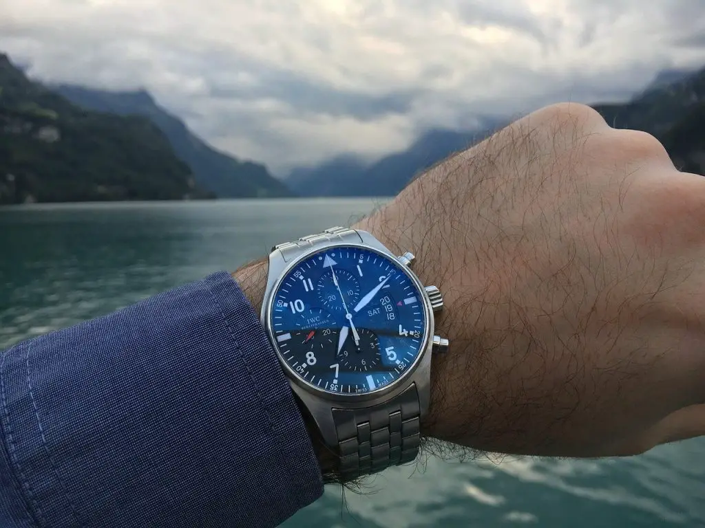 A blue-faced IWC watch worn by a man in a navy-blue suit. The background is that of a bay with mountains in the background.